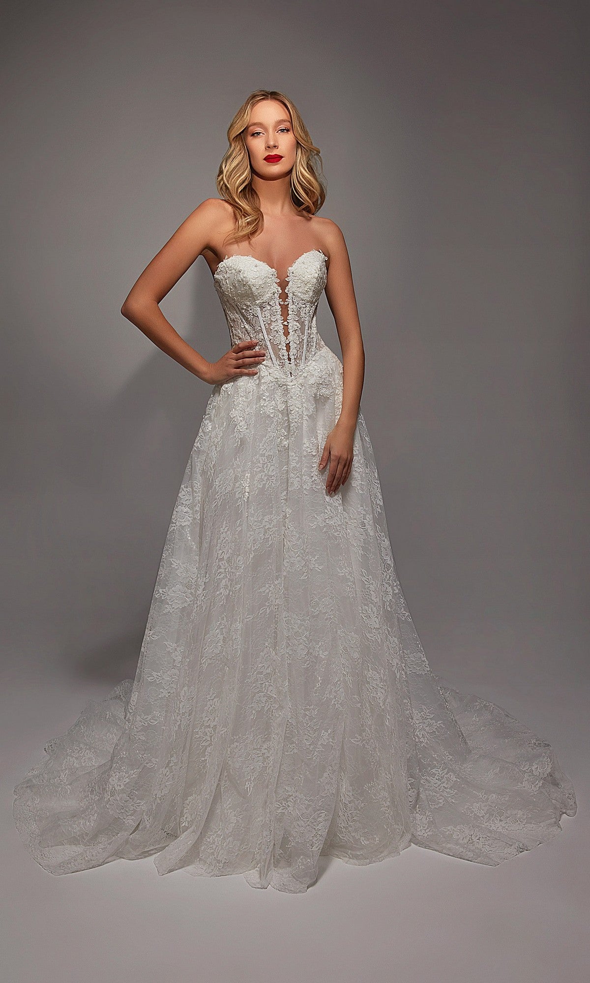 A woman wears the Alyce 7154 long white dress in ivory/sand, featuring a strapless lace design with a sheer corset bodice and flowing skirt, standing before a plain gray backdrop.